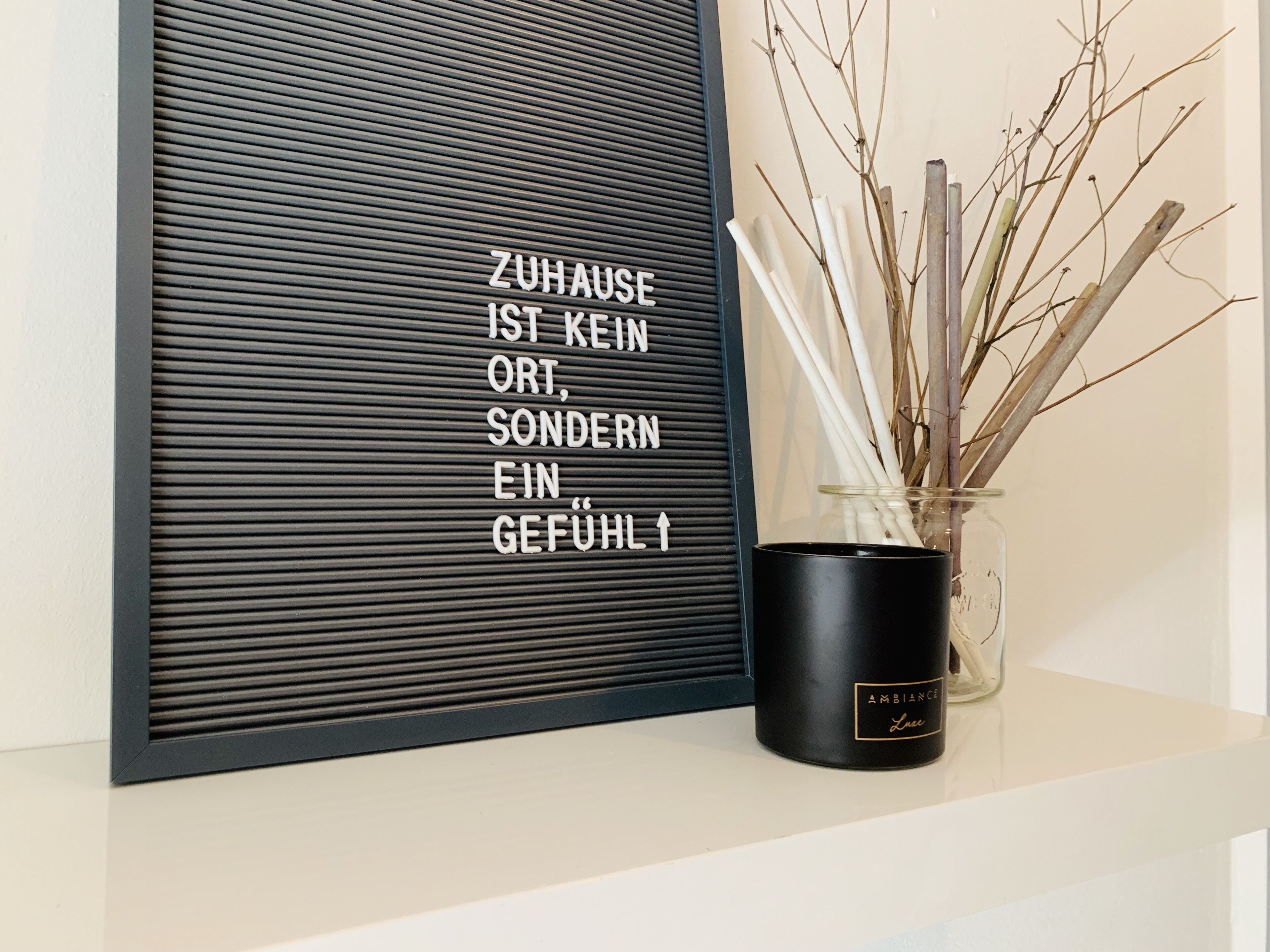 Ein gerahmter Spruch in deutscher Sprache, eine schwarze Vase mit weißen Ästen und ein schwarzer Behälter mit der Aufschrift 'AMBIENTE Luxe'.