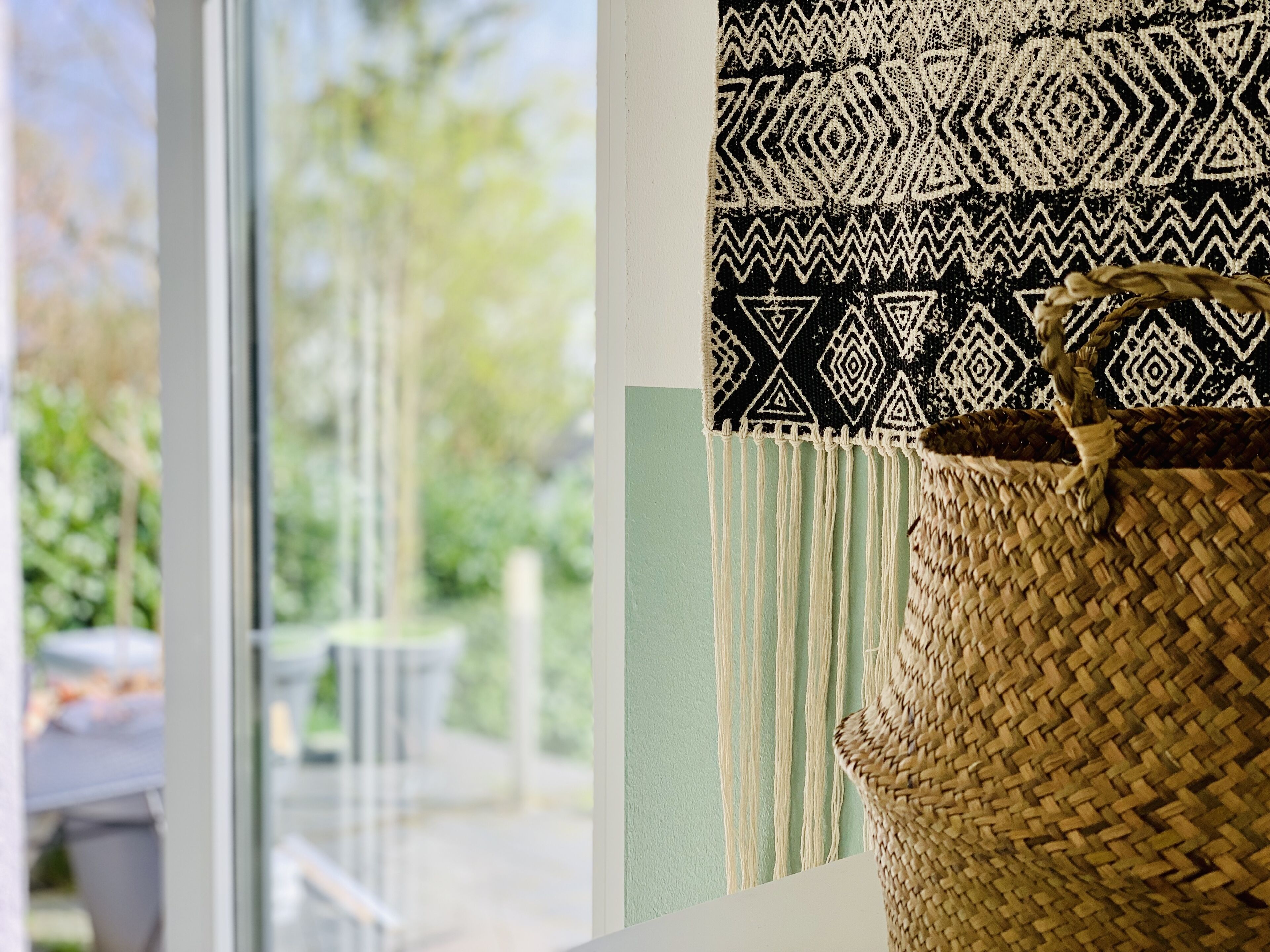 A woven basket with a handle, a patterned fabric, and a view of a green outdoor area through a window.