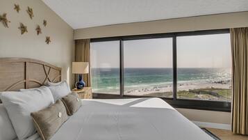 Hotelkamer met een groot bed, een nachtkastje met een lamp en uitzicht op het strand en de zee door een groot raam.