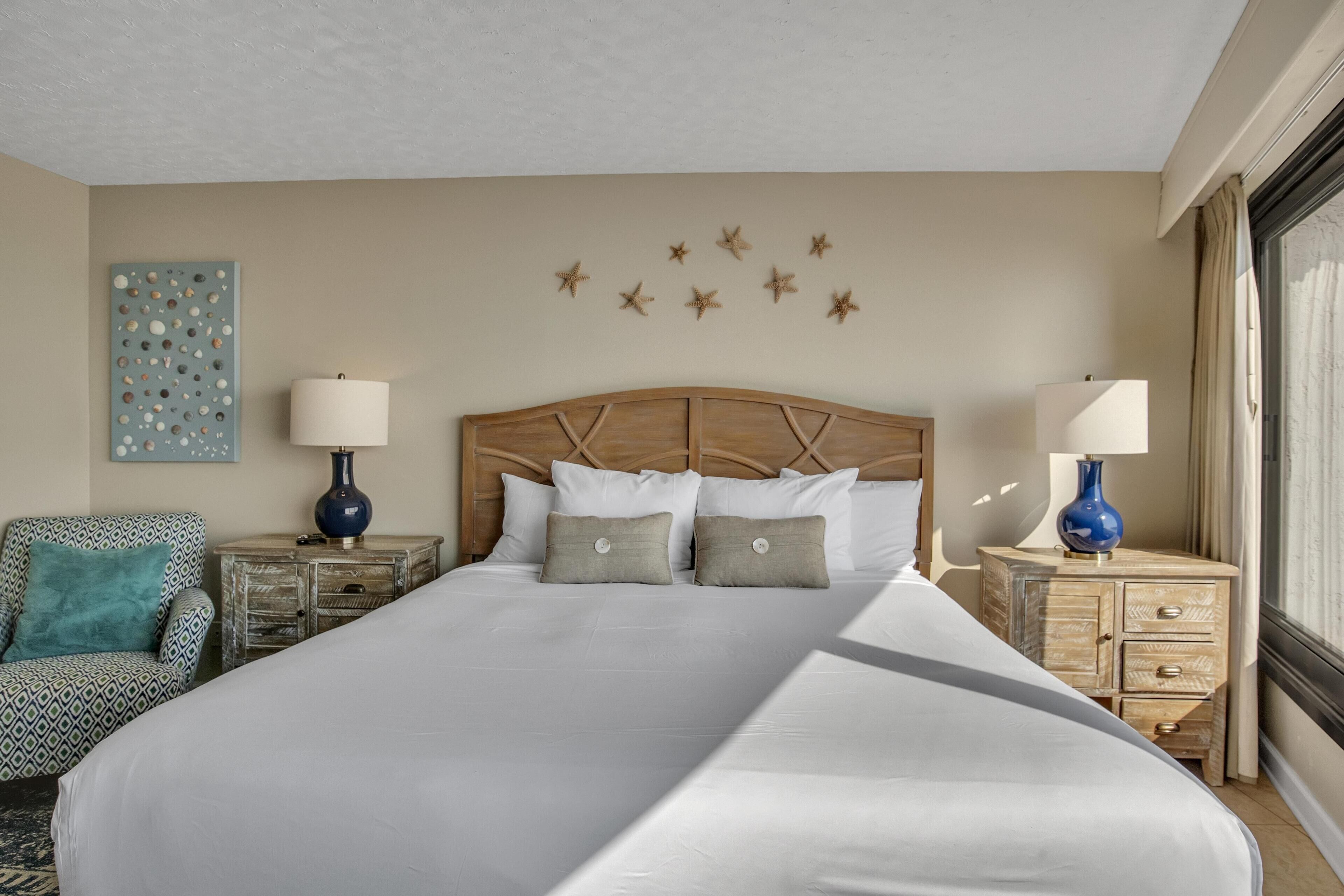A neatly made bed with a wooden headboard, two bedside tables with lamps, a patterned armchair, and a wall art piece.
