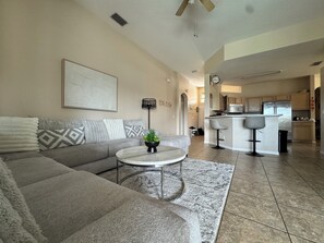 A living room with a grey sofa, a coffee table, and a plant. There is a kitchen area with a bar and a refrigerator. - 4 Bedroom Executive Villa with Pool & Spa, Games Room, Wifi and Large Garden
(Davenport)