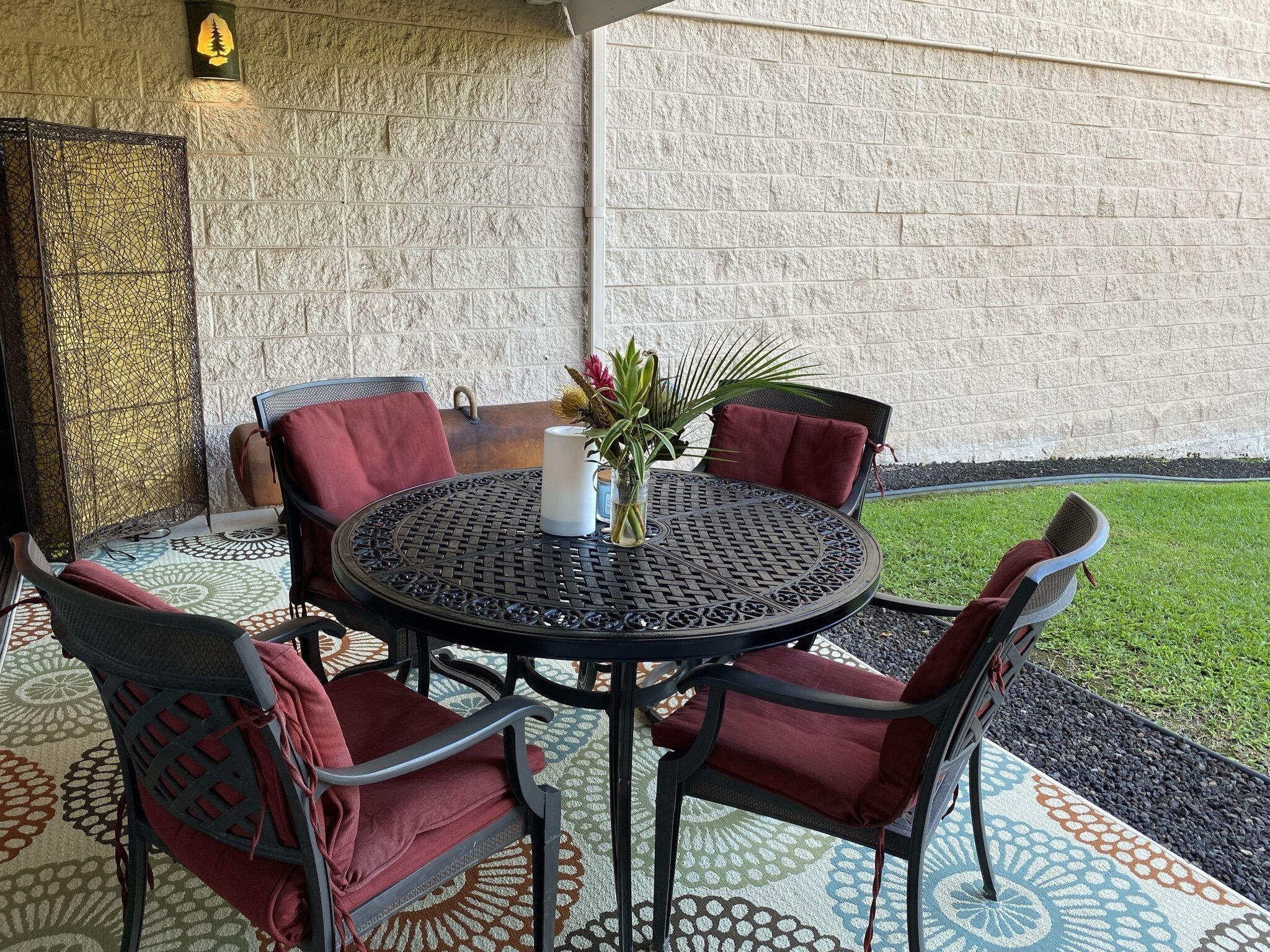 Ein runder Metalltisch mit vier Stühlen, jeder mit einem roten Kissen. Auf dem Tisch steht eine weiße Vase mit Blumen. Im Hintergrund ist eine Steinsichtmauer und eine Rasenfläche zu sehen.