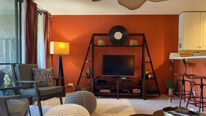 Una sala de estar con televisor sobre un mueble bajo, un sofá, una mesa de centro y una zona de cocina con taburetes.