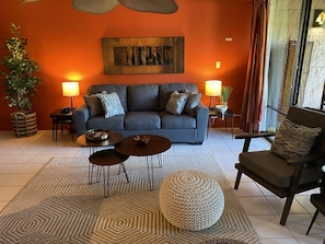 Una sala de estar moderna con un sofá gris, dos mesitas de noche con lámparas, una mesa de centro y un otoman redondos, además de una planta en maceta.