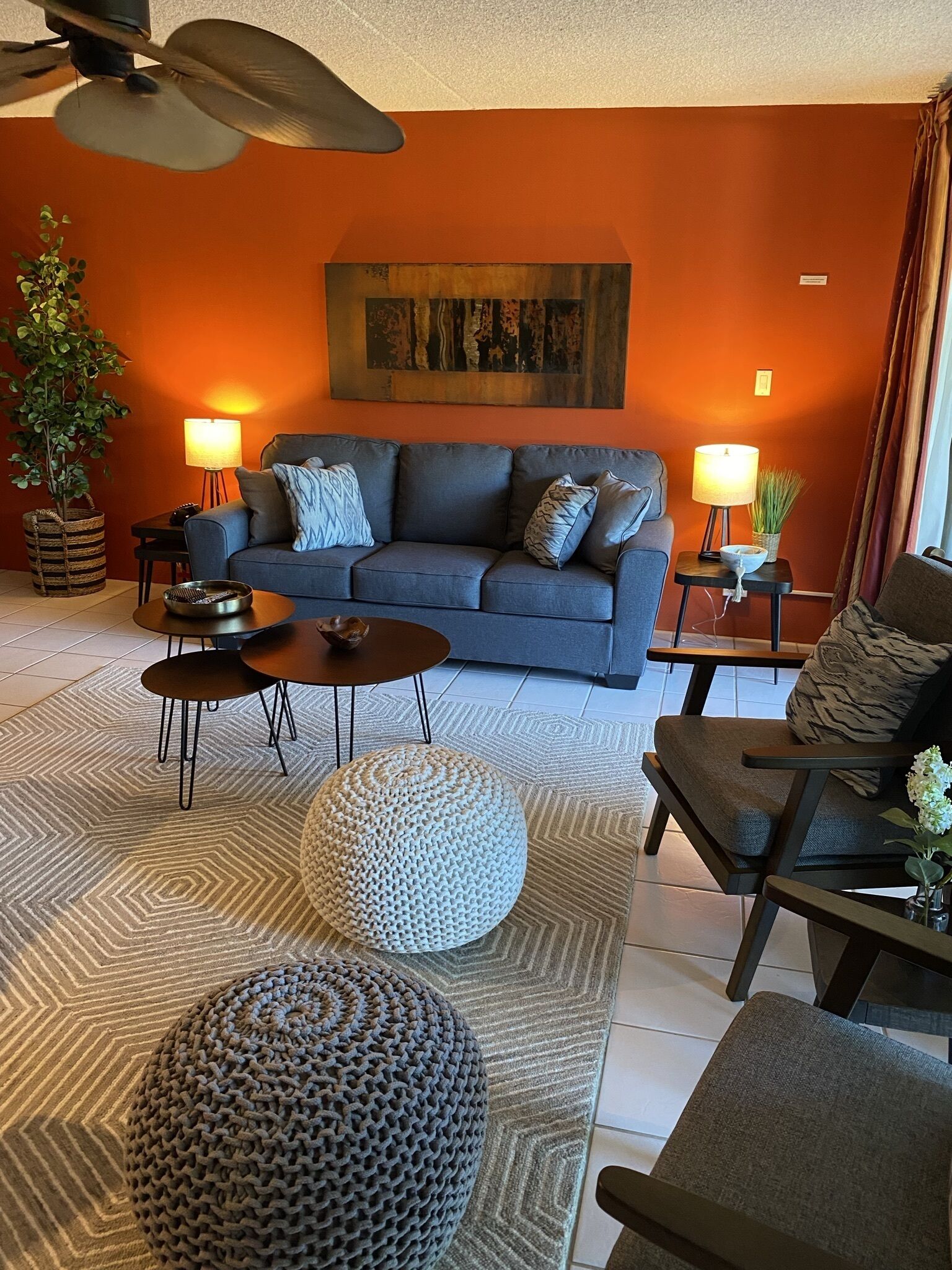 A modern living room with a blue sofa, two side tables with lamps, a round ottoman, and a ceiling fan.