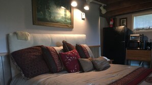 Een net opgemaakt bed met meerdere kussens, een schilderij aan de muur, en een magnetron en koelkast op de achtergrond.
