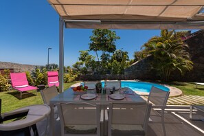 Um pátio externo com mesa posta para refeições, rodeado por cadeiras, piscina e palmeiras.
