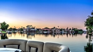 A boat with a cushioned seating area and a small table, docked near a calm body of water with buildings and palm trees in the background.