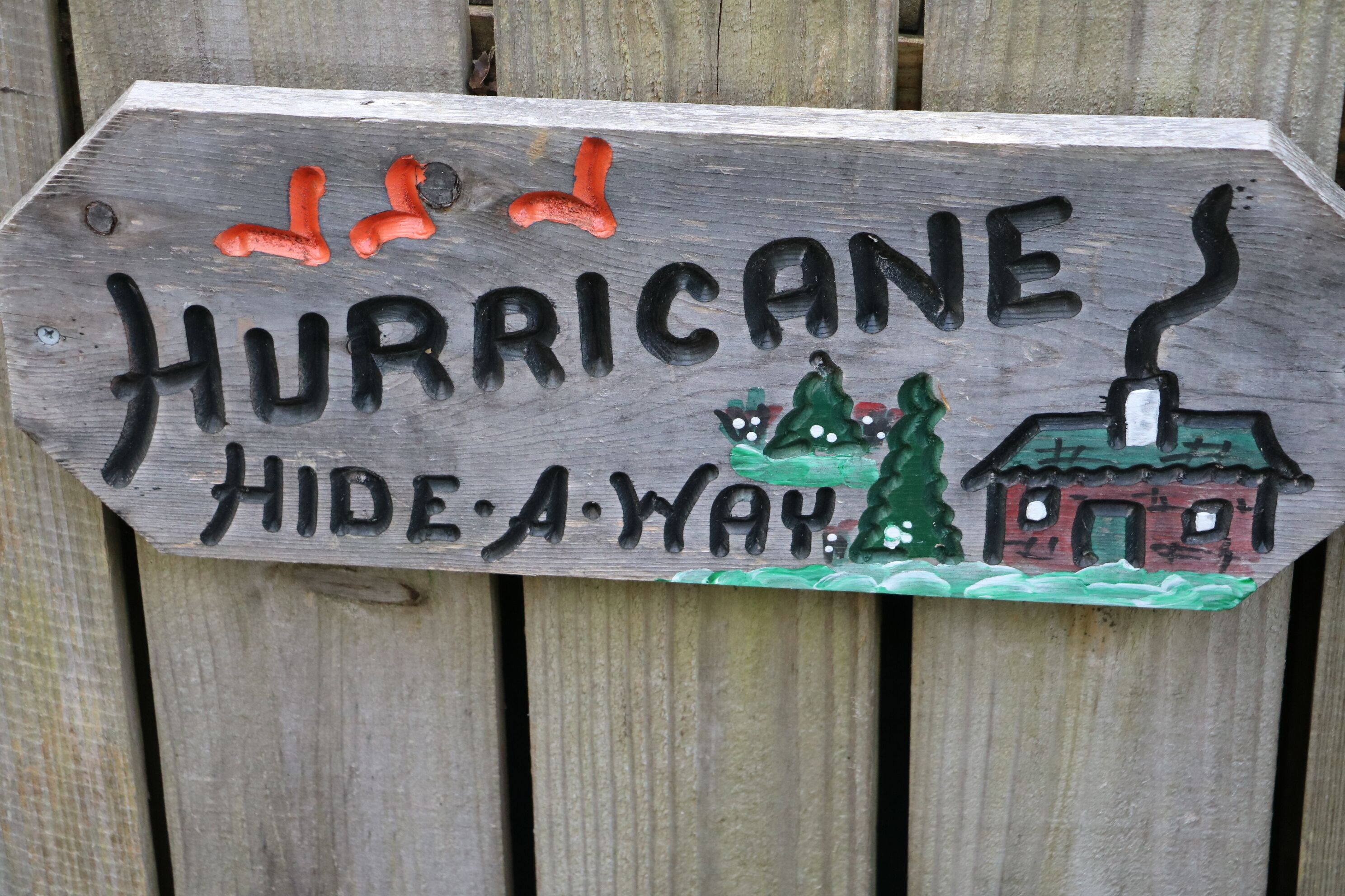 A wooden sign with the words "HURRICANE HIDE-A-WAY" and illustrations of a house, trees, and a chimney.