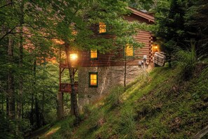 Una cabaña de troncos con terraza de madera y pared de piedra, rodeada de un bosque frondoso.