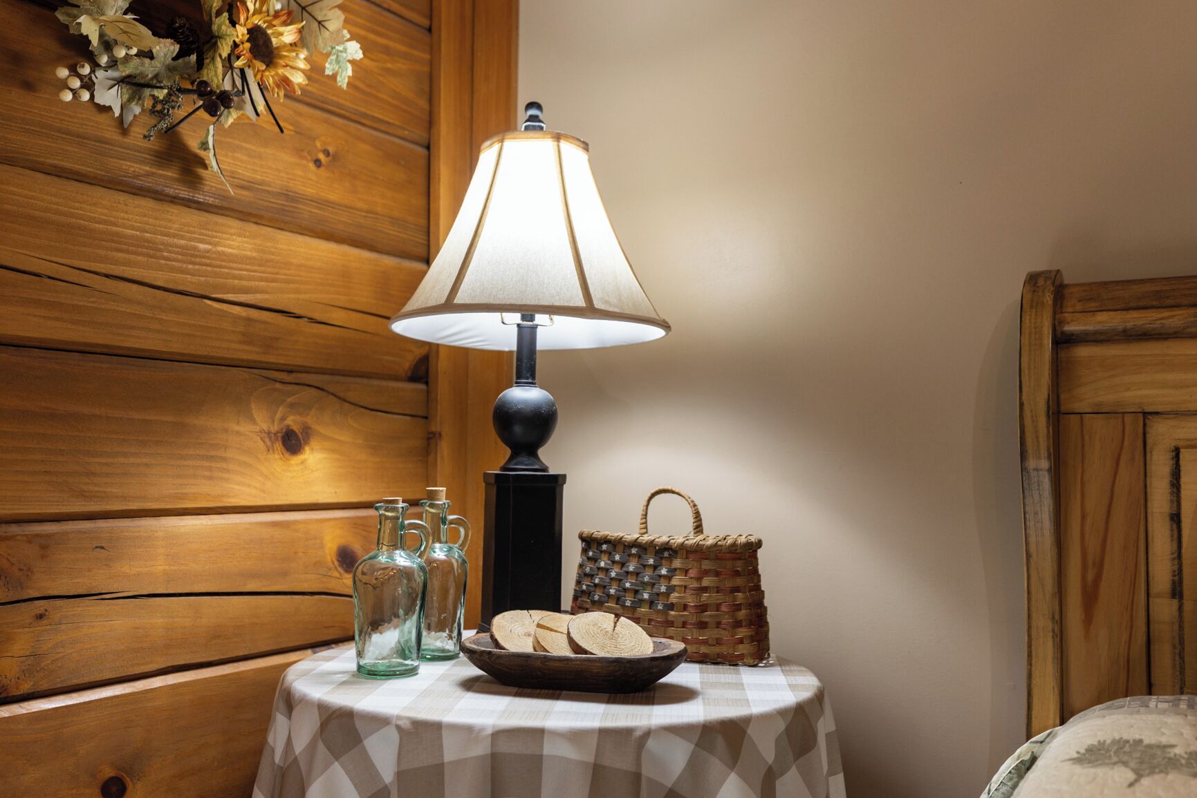 A wooden wall, a table with a lamp, glass bottles, a basket, and a checkered tablecloth.