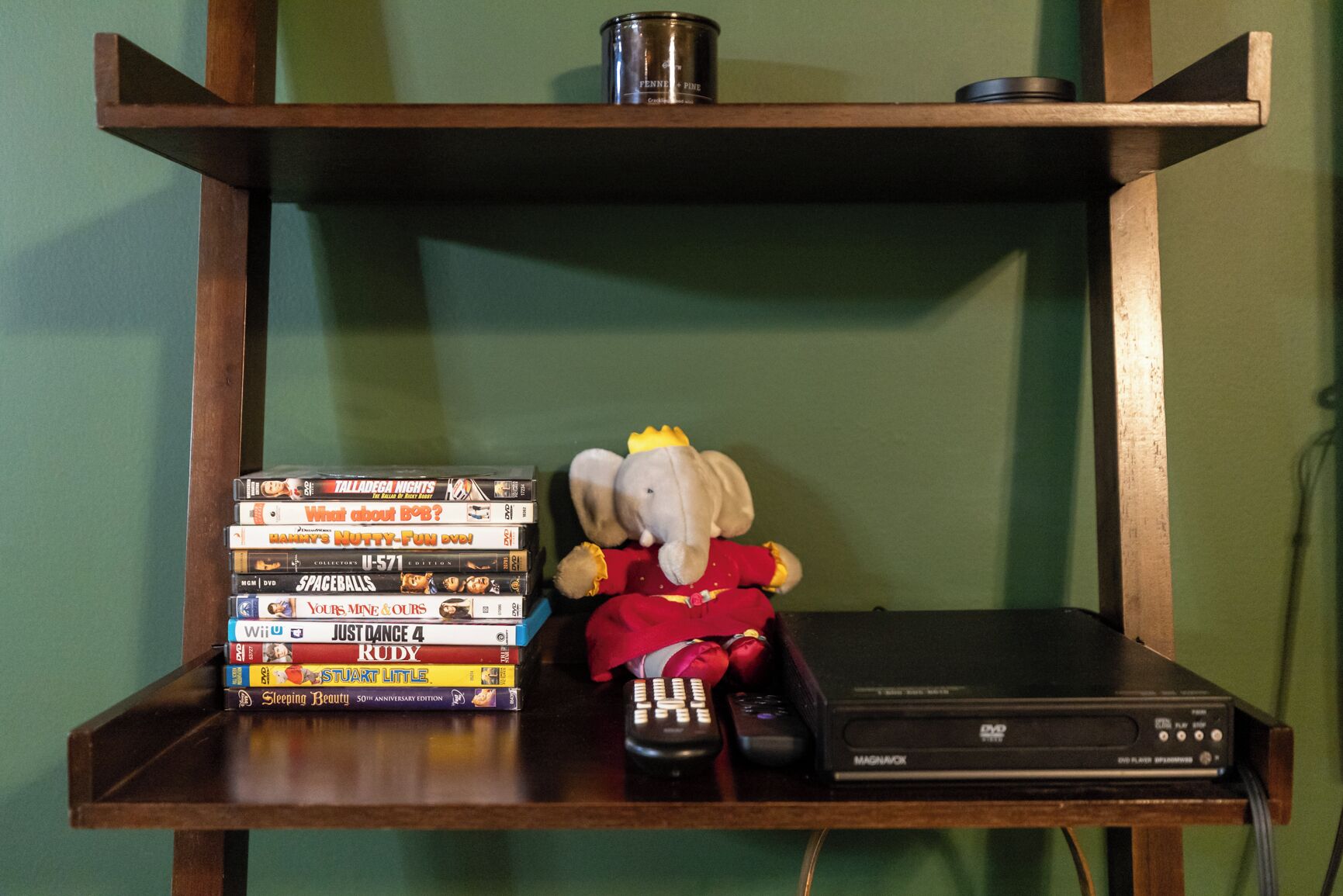 Uma prateleira de madeira com uma pilha de DVDs, um brinquedo de pelúcia, um controle remoto e um aparelho de DVD.