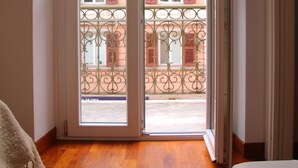Quarto com piso de madeira, um sofá branco e vista para uma varanda com persianas vermelhas e grade de ferro forjado.