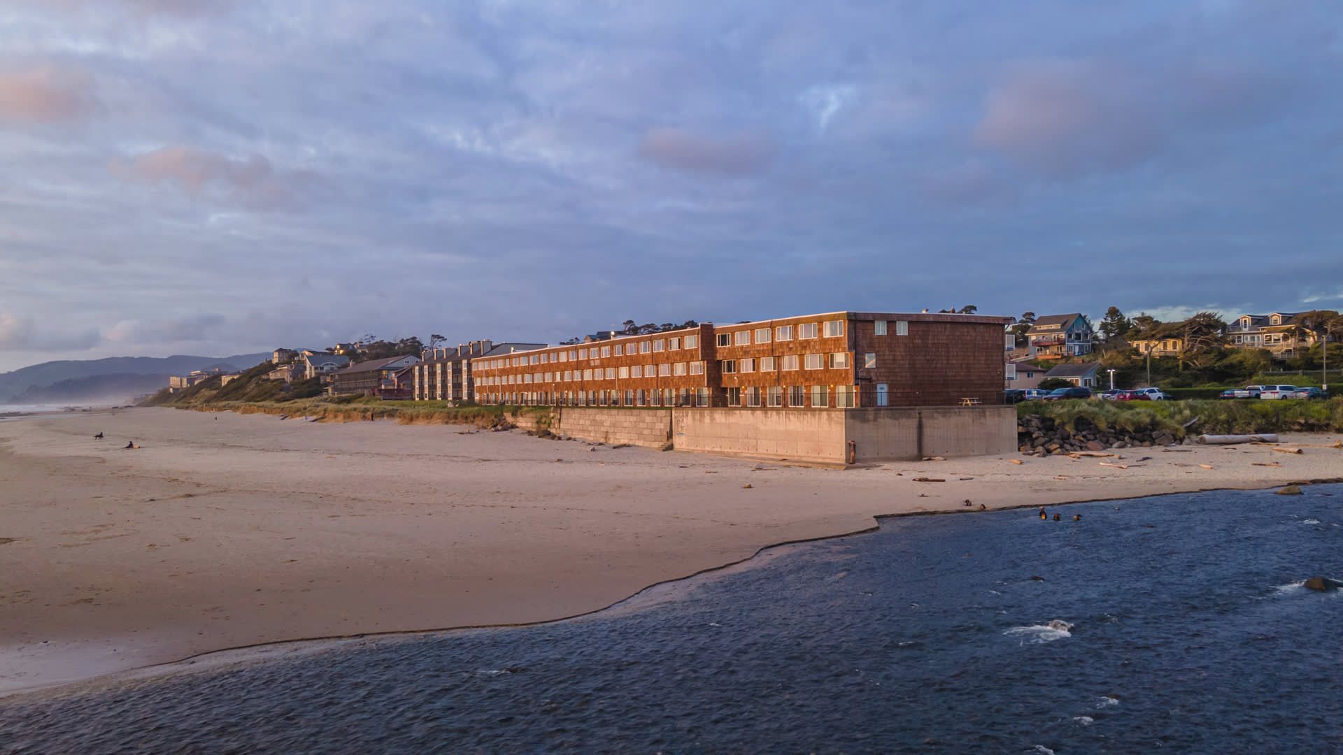 Un bâtiment situé sur la côte, doté d’un toit plat et de nombreuses fenêtres, et qui est adjacent à une plage de sable et à une étendue d’eau.