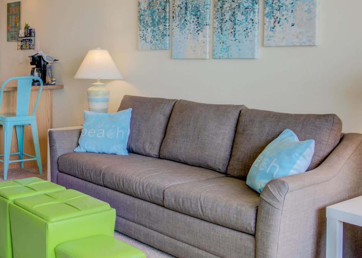 A living room with a grey sofa, two blue pillows, a green ottoman, a white lamp, and abstract art on the wall.