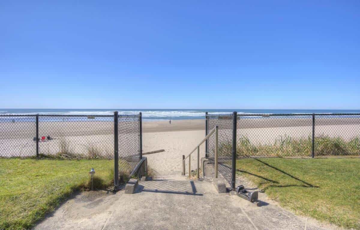 A concrete pathway leading to a beach with a fence on one side and a grassy area on the other.