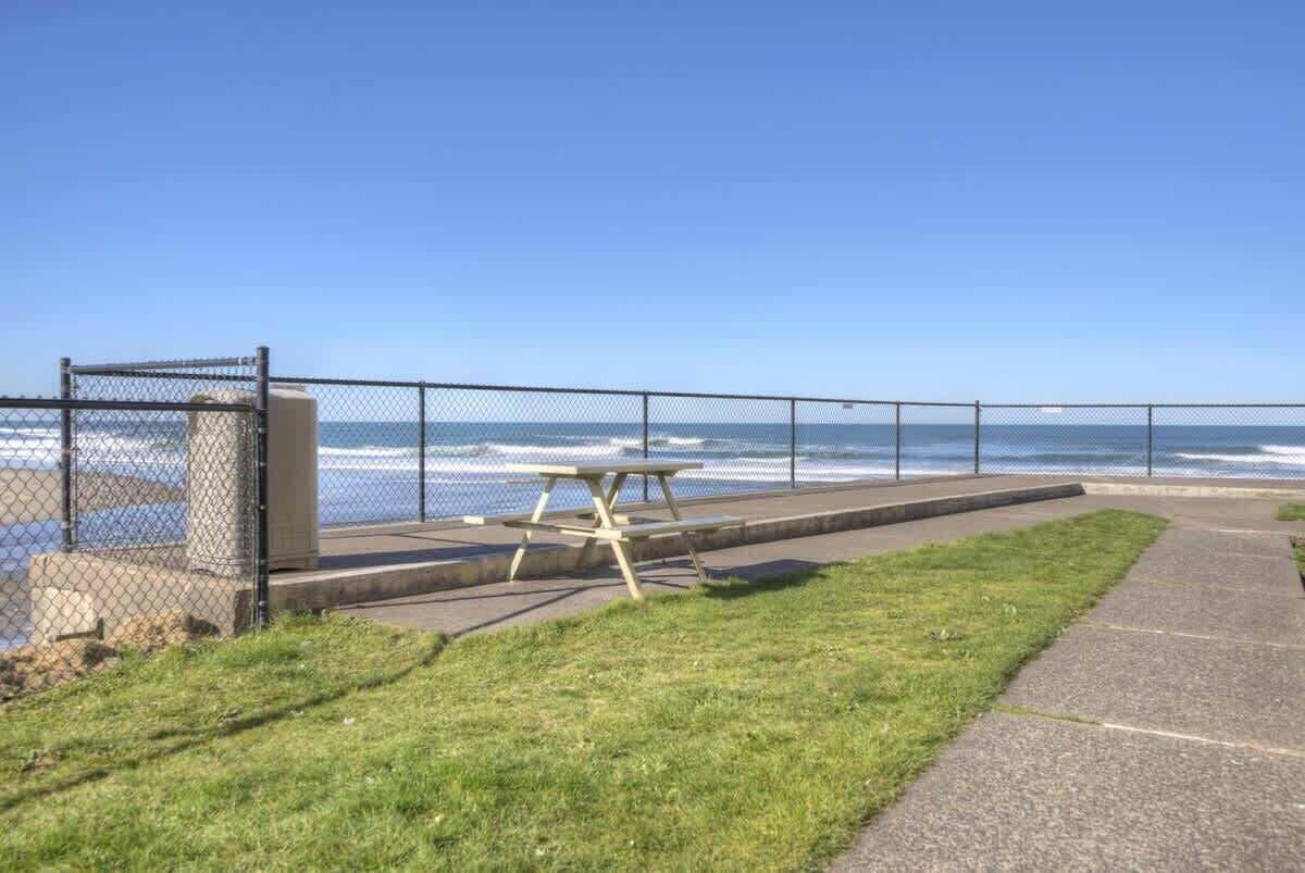 A picnic table and bench by the ocean, with a fence and grassy area in the foreground.