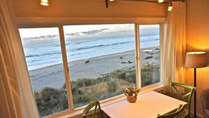 Una habitación con un ventanal grande con vista a una playa, una mesa con sillas y una lámpara.