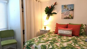 A bedroom with a bed, a nightstand, a lamp, a chair, and a painting of a beach scene.