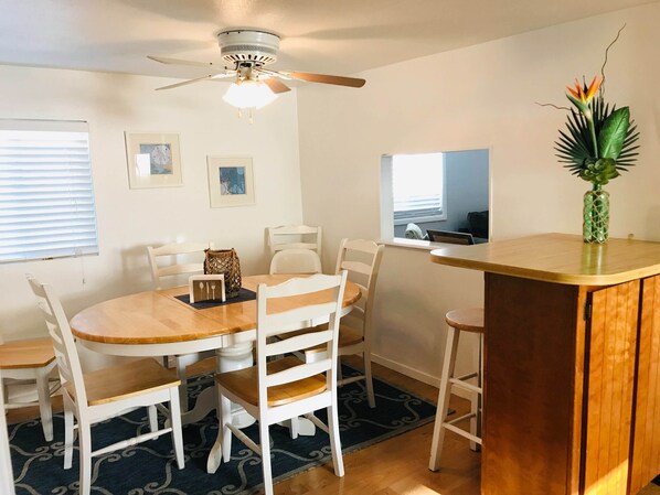 Un área de comedor con una mesa redonda de madera, sillas blancas, un ventilador de techo y una pequeña planta decorativa sobre un mostrador de madera.