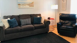 A living room with a dark brown sofa, a black leather chair, a painting of a beach scene, a floor lamp, and a clock on the wall.