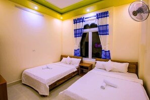A hotel room with two single beds, a window with blue curtains, a standing fan, and a wooden nightstand. - Gia Thinh Hotel (Ly Son)