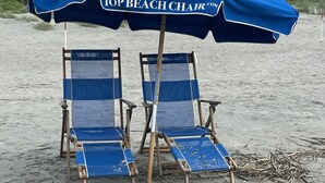 Zwei Liegestühle unter einem blauen Sonnenschirm mit weißem Logo, an einem sandigen Strand mit etwas Treibgut.