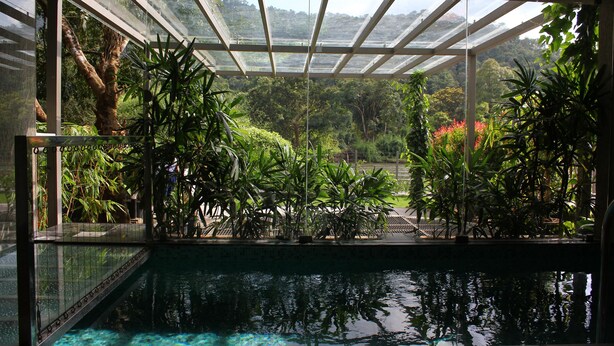 A pool area with a glass enclosure, surrounded by lush tropical plants and a view of a forested landscape. - Niraamaya Retreats Samroha Athirappilly (Chalakudy)