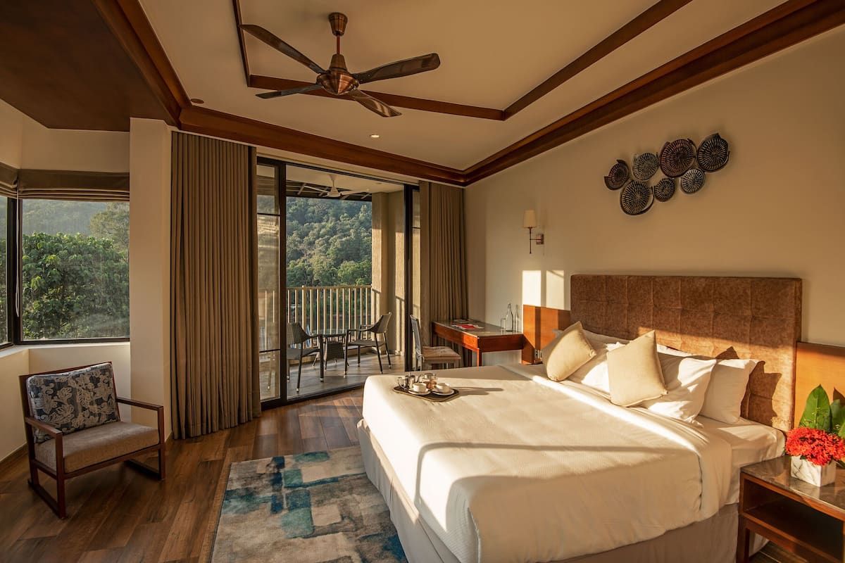 A hotel room with a large bed, a desk, a chair, and a balcony with a view of the forest.