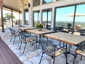 Een terras buiten met stenen vloer, houten tafels, metalen stoelen en een plant in pot.