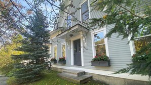 Une maison à deux étages avec un porche à l’avant, un petit escalier et un jardin devant la maison, avec un arbre et des jardinières.