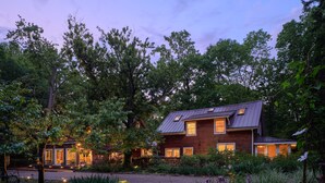 A two-story wooden house with a sloped roof, surrounded by lush greenery and trees.