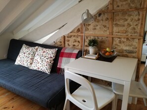 A cozy loft bedroom with a dark blue sofa, a white desk, and a white chair. There is a small potted plant on the desk and a fruit bowl on the wall.