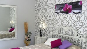 Ein Schlafzimmer mit einem Bett, einem Nachttisch, einem Spiegel, einem Wandbild und einer Vase mit Blumen.