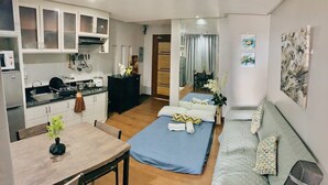 Una habitación de hotel compacta con kitchenette, una cama con ropa de cama azul, un sofá con cojines estampados y obras de arte en las paredes.