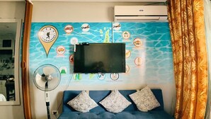Habitación de hotel con cama, televisor de pantalla plana montado en la pared, ventilador de pie y una pared decorativa con varios iconos.