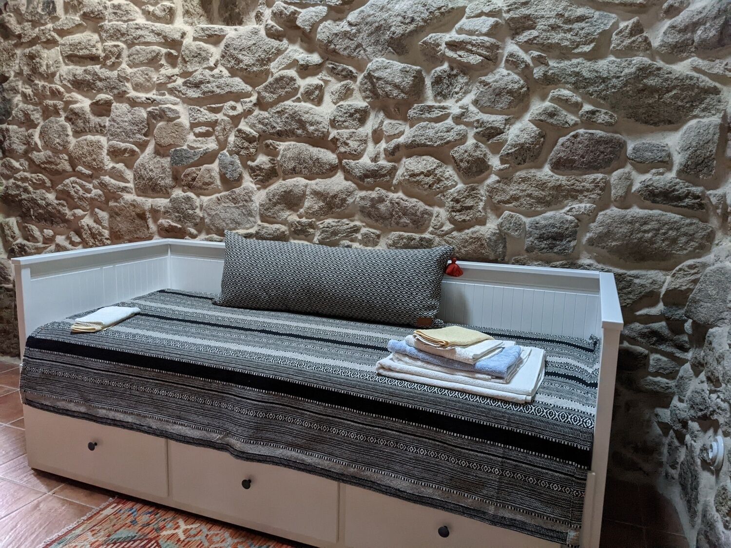 A small bed with a patterned blanket and pillows against a stone wall.