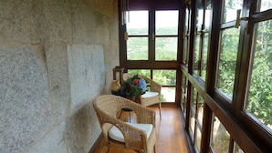 A room with a stone wall, wooden floor, wicker chairs, a small table with a vase and flowers, and large windows with a view of greenery.