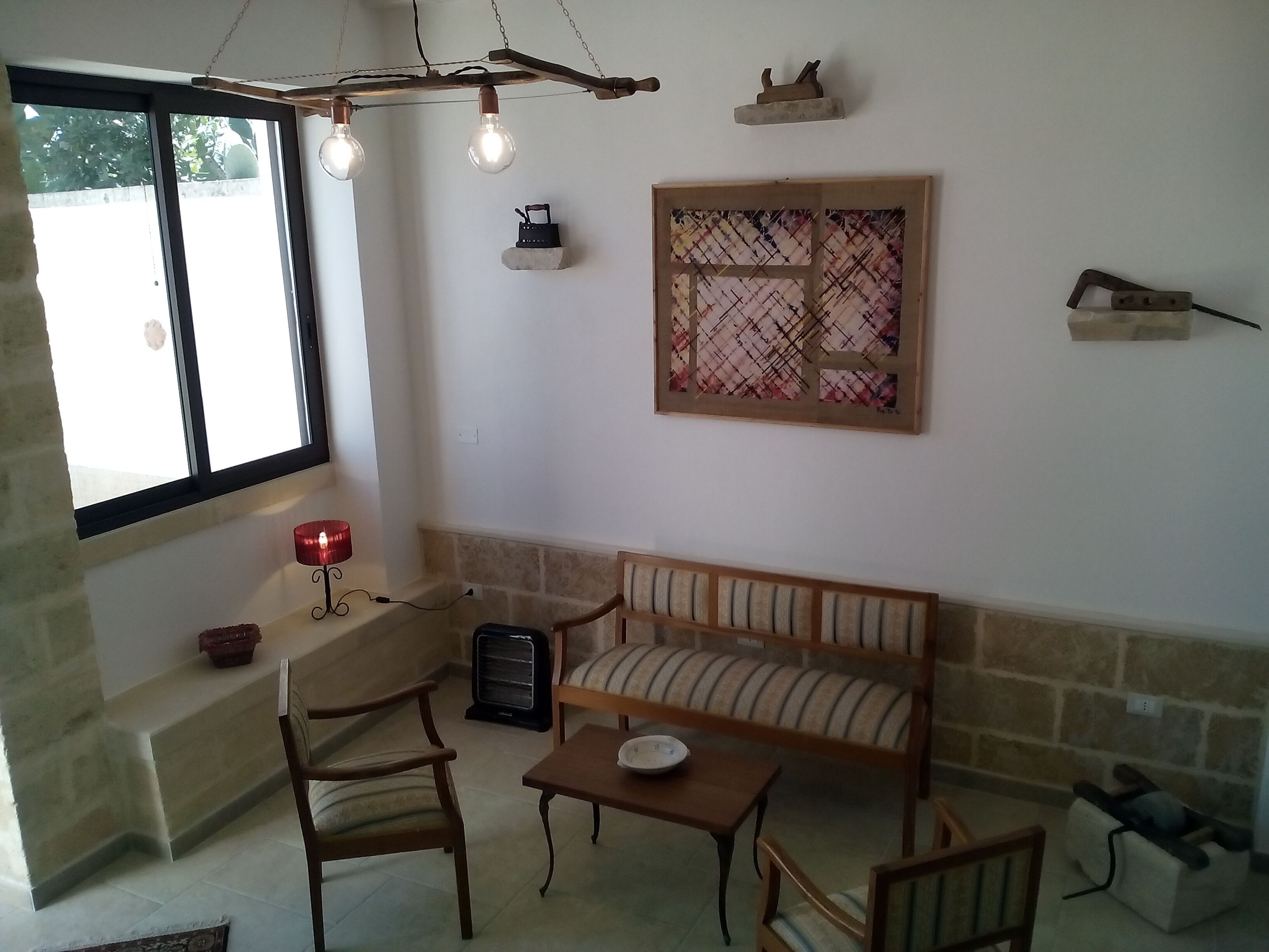 Uma sala de estar acolhedora com um sofá, duas poltronas, uma mesinha, um lustre decorativo, um quadro na parede e uma janela com vista.