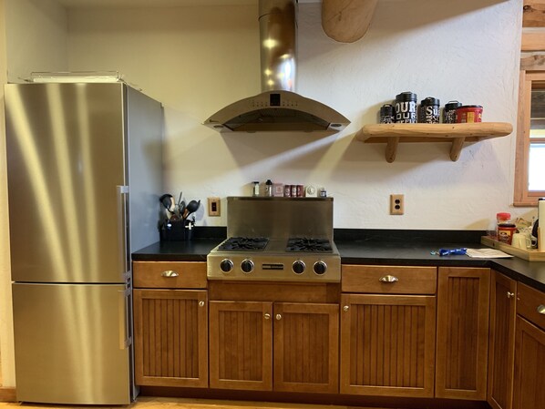 A kitchen with a stainless steel refrigerator, a gas stove, wooden cabinets, and a wooden shelf with various containers.