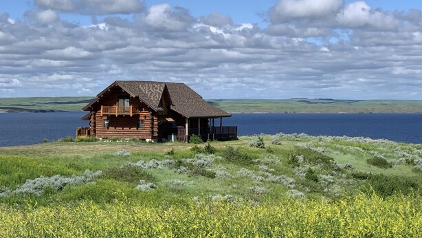 Une cabane en rondins dotée d’un balcon, située sur une colline verdoyante et offrant une vue sur une étendue d’eau.