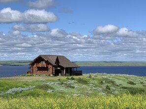 A log cabin with a balcony, situated on a grassy hill overlooking a body of water.
