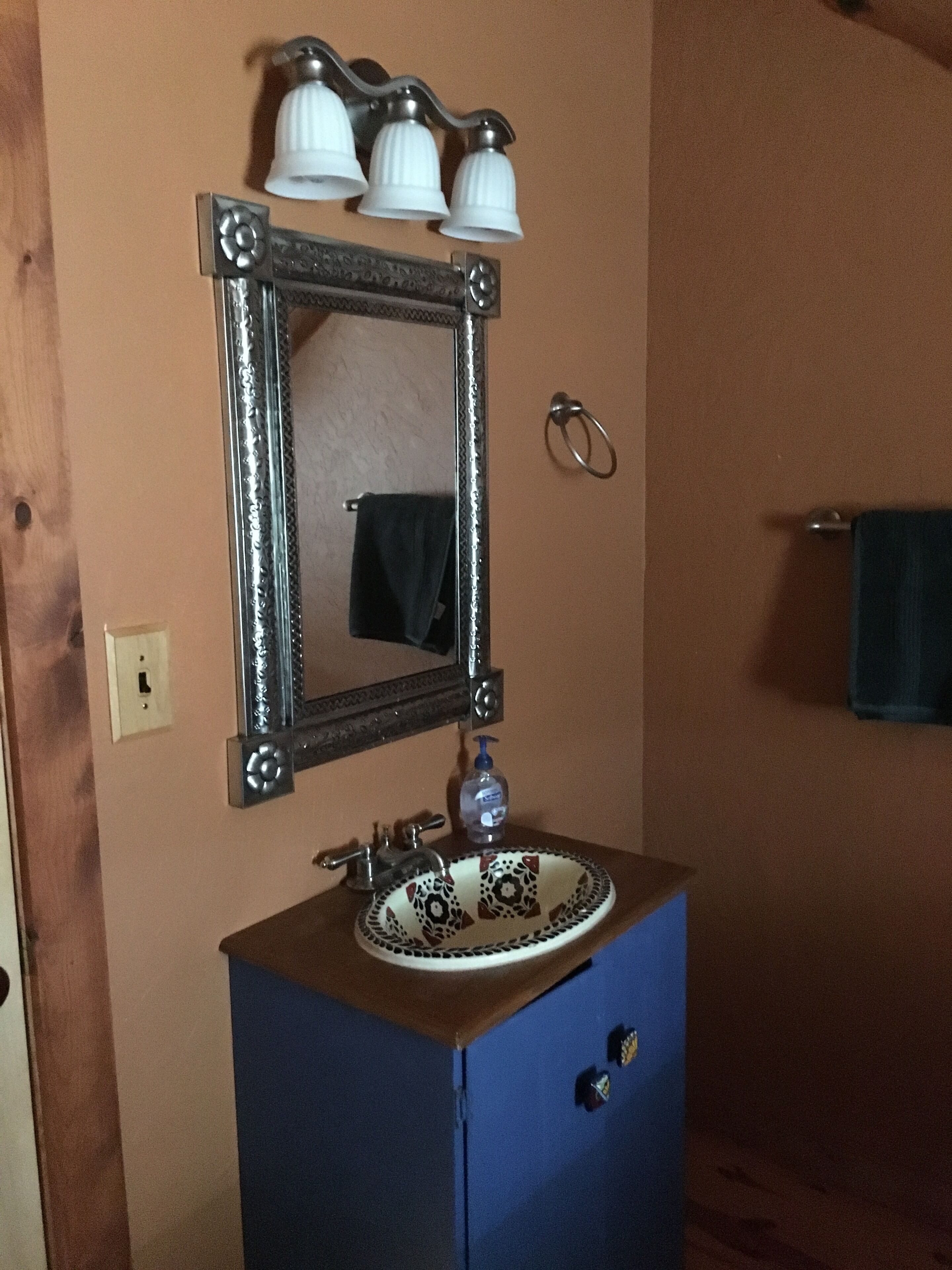 A bathroom with a blue vanity, a mirror with three lights, and a towel ring on the wall.