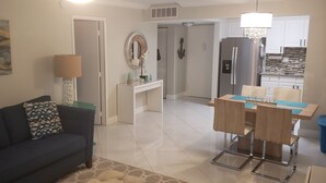 Un saló modern amb un sofà fosc, una taula de centre i una làmpada. Hi ha una zona de menjador amb una taula i cadires. Es veu una cuina al fons amb un frigorífic i un rentaplat.