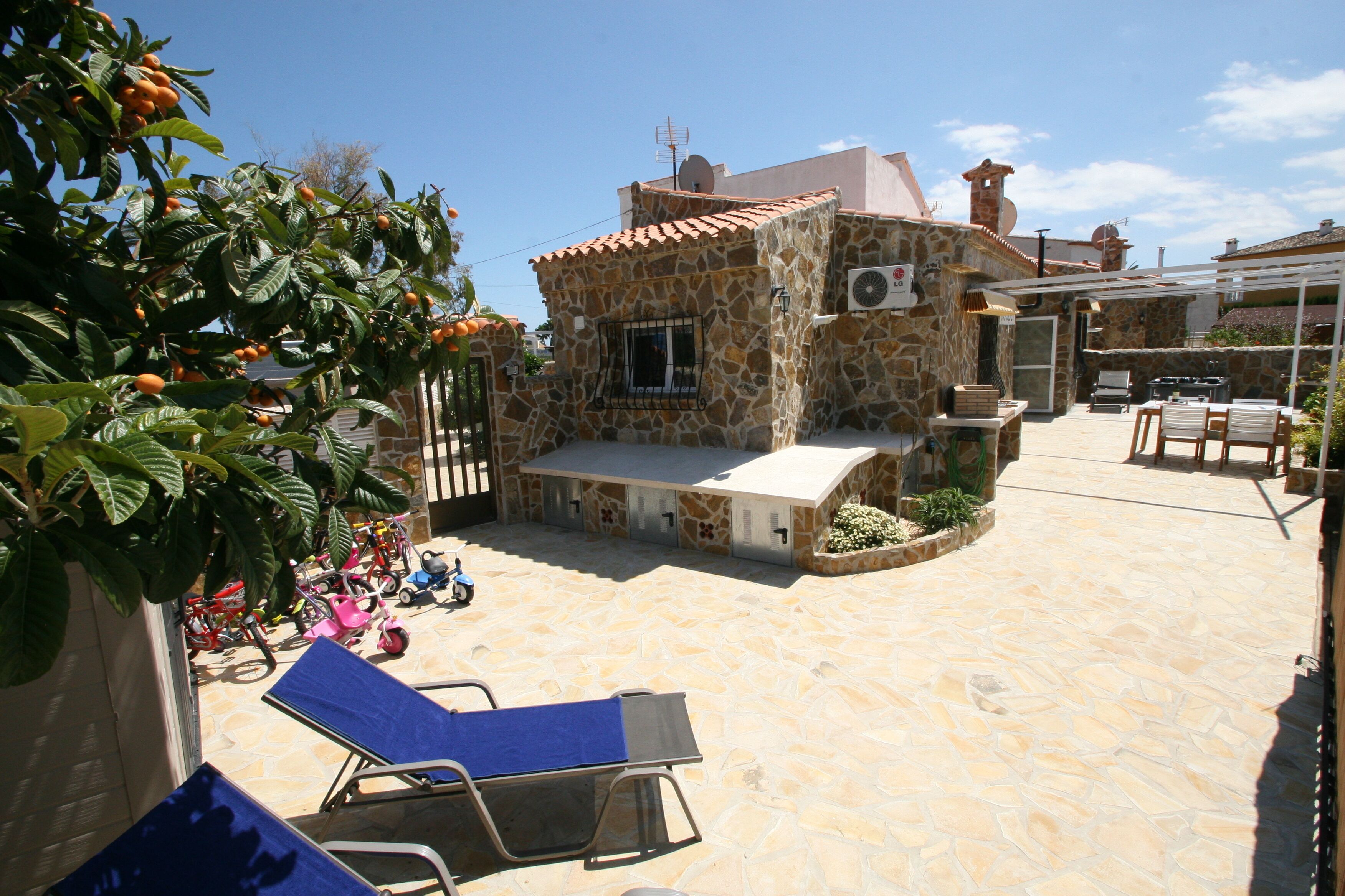 A stone building with a tiled roof, a small patio area with lounge chairs, and a garden with fruit trees.