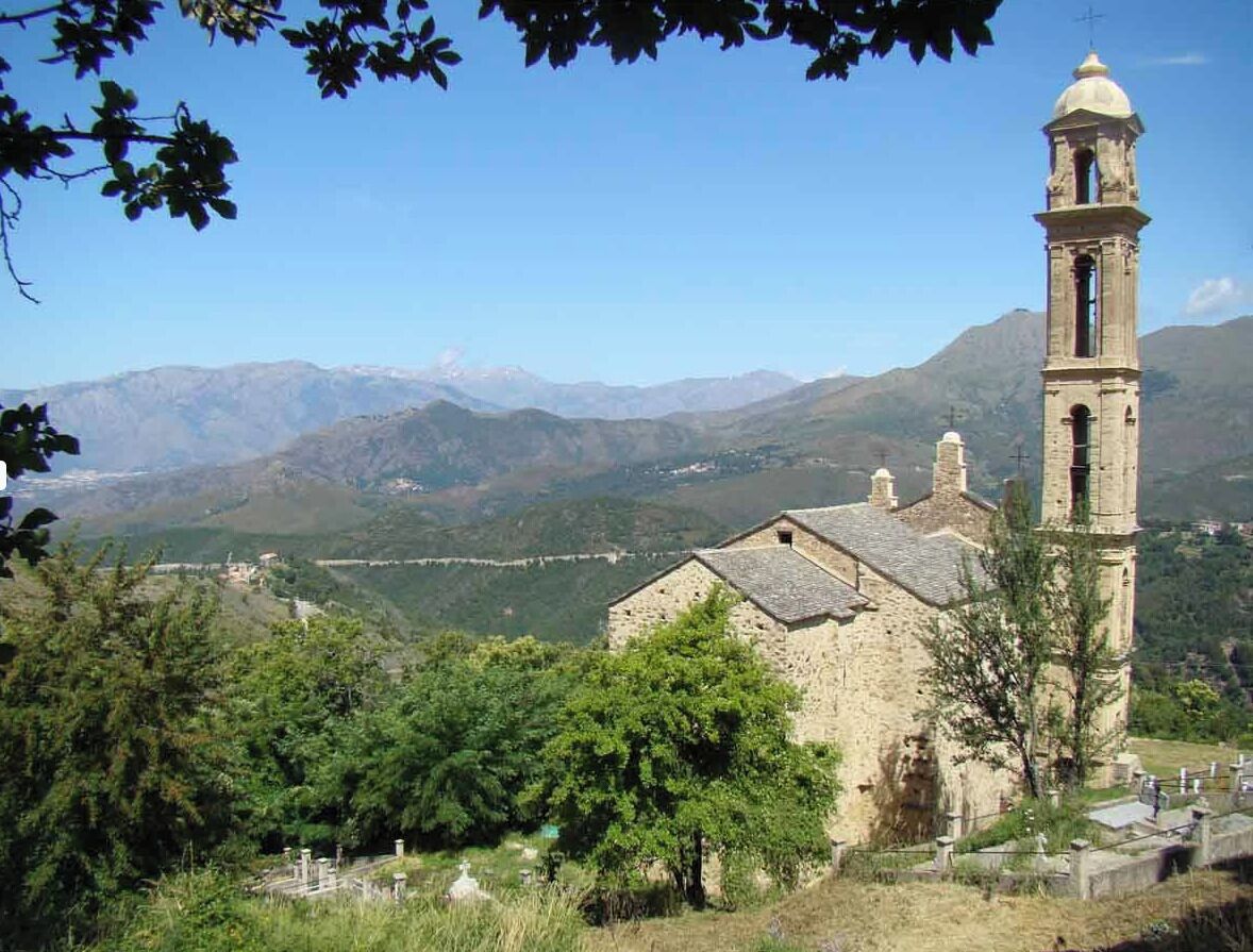A stone church with a bell tower, surrounded by trees and mountains.