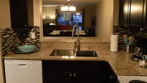 A kitchen with a granite countertop, stainless steel sink, and a dishwasher. There is a dining area visible through the window. - Top floor Ocean View Cocoa Beach Condo at the Spanish Main (Cocoa Beach)