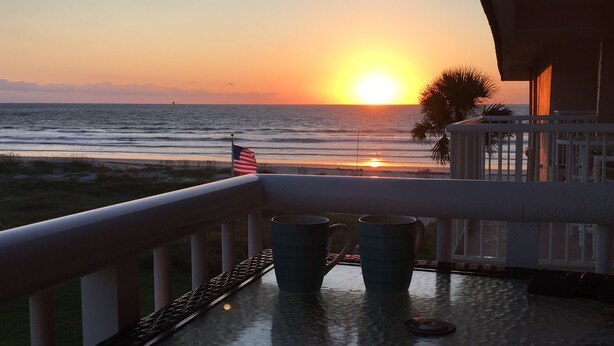 A balcony with a view of the sunset over the ocean, a palm tree, and two mugs on the table. - Top floor Ocean View Cocoa Beach Condo at the Spanish Main (Cocoa Beach)