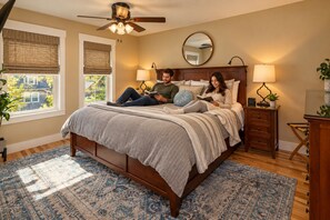 A bedroom with a large bed, two bedside tables, a ceiling fan, and a view of the outside.