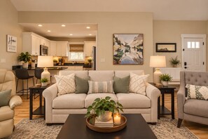 A living room with a sofa, armchairs, a coffee table with a tray of books and candles, a kitchen in the background, and a painting on the wall.